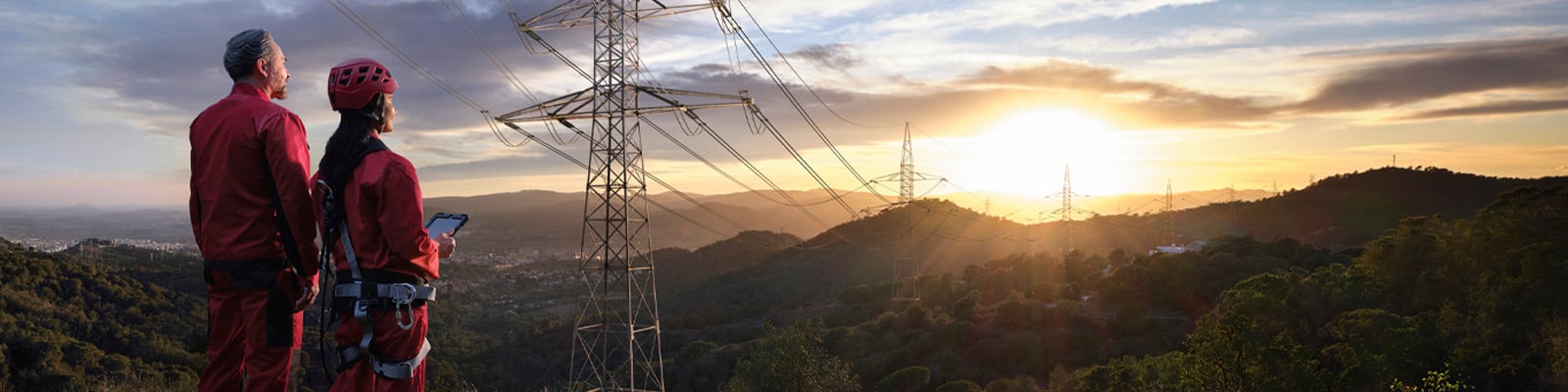 Two workers in red boiler suits watch the sun set over pylons